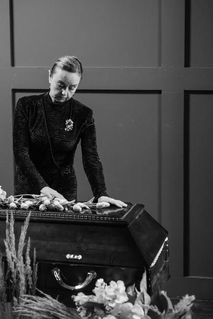 A woman dressed in a dark, textured outfit with a brooch on her chest stands solemnly over a closed casket, gently placing a floral arrangement of white roses and greenery on top. The casket is dark w