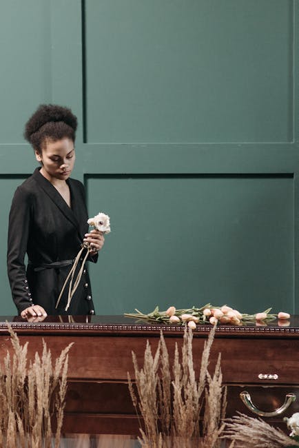A young woman with curly dark hair, dressed in a black blazer, is standing beside a wooden casket inside a funeral setting with muted green walls. She is holding a single white flower with a delicate,