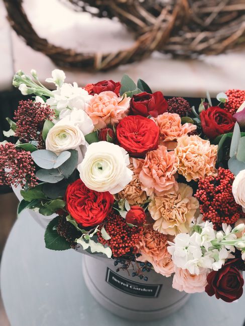 A vibrant floral arrangement in a round white hatbox featuring red, white, peach, and blush pink flowers, including roses, ranunculus, and carnations, complemented by dark red hypericum berries and so