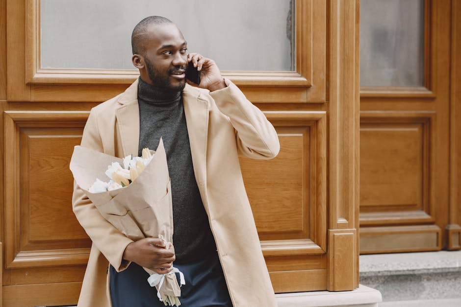 A man with a dark complexion, neatly groomed beard, and short hair is standing outdoors against a wooden-paneled wall. He is holding a bouquet of white flowers wrapped in brown paper in his left hand,
