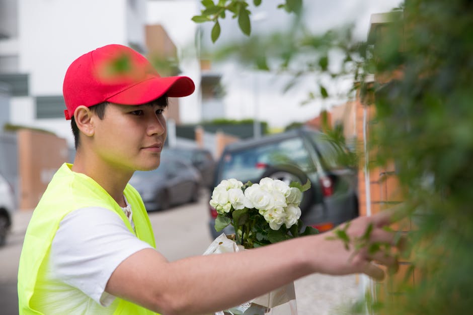 A young delivery person wearing a red cap and a fluorescent yellow vest is seen reaching out to hang a floral gift on a green shrub outside a residential area. The bouquet features white hydrangeas an