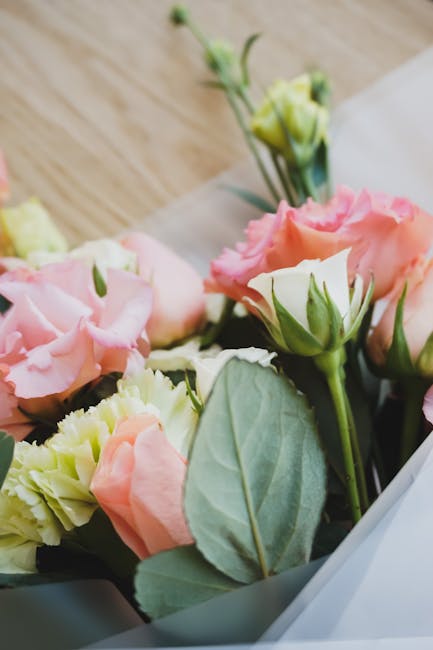 Close-up image of a floral arrangement featuring soft pink and cream roses with ruffled petals, complemented by green leaves and buds. The bouquet is wrapped in pale grey paper and is positioned on a 
